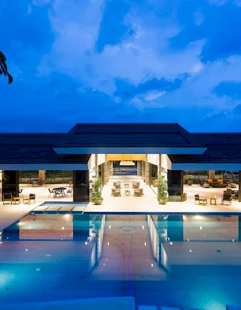 An illuminated luxury building features an outdoor pool with modern seating areas around, set against a twilight sky.