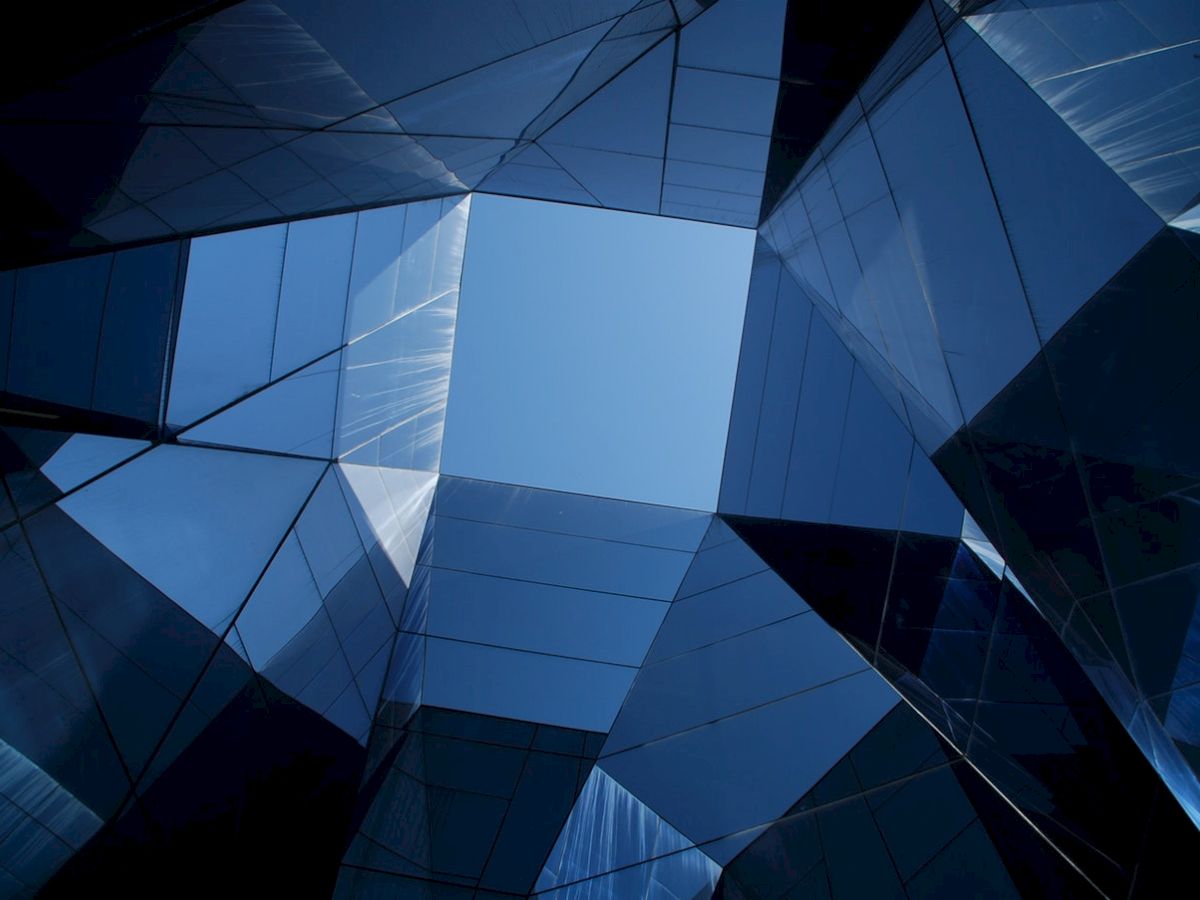 Abstract view from the bottom of a modern glass building, looking up at a patch of blue sky framed by angular glass and steel structures.