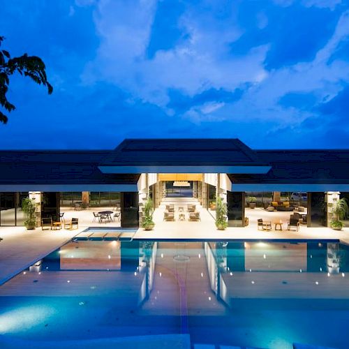 The image shows a luxurious outdoor swimming pool area at dusk, with a modern building and open seating areas in the background.