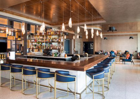 The image shows a modern bar with high chairs, hanging lights, and a well-stocked liquor shelf. The seating area has sofas and tables.