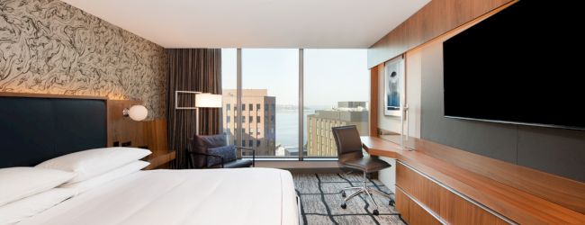 A modern hotel room with a large bed, wall art, desk, chair, and large TV. The window offers a view of city buildings and possibly water.