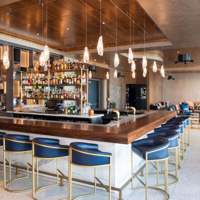 A modern bar with a wooden countertop, blue bar stools, a well-stocked liquor shelf, pendant lights, and seating areas in the background.