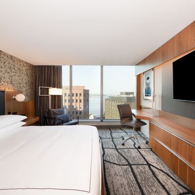 A modern hotel room with a bed, desk, chair, large TV, and a window offering a city view. The decor features wooden accents and a patterned wall.