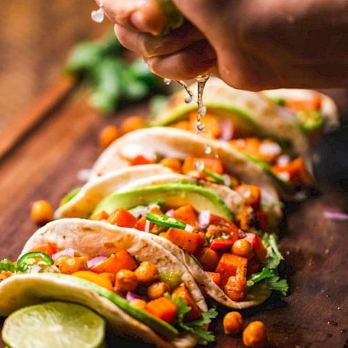 The image shows a hand squeezing lime over a row of chickpea and vegetable tacos garnished with avocado and cilantro on a wooden board.