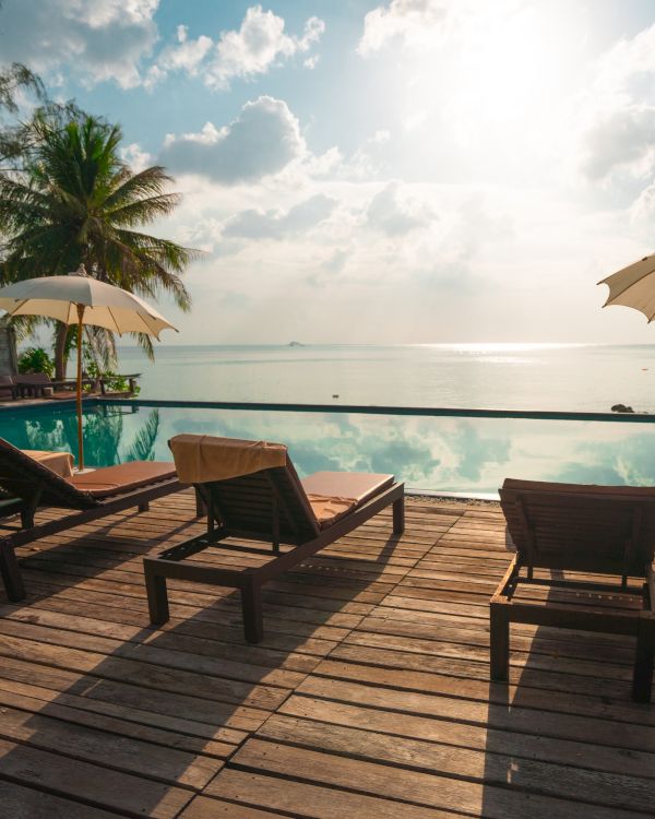 Sun loungers and umbrellas by a serene swimming pool, overlooking the ocean with a bright sun and some clouds in the sky.
