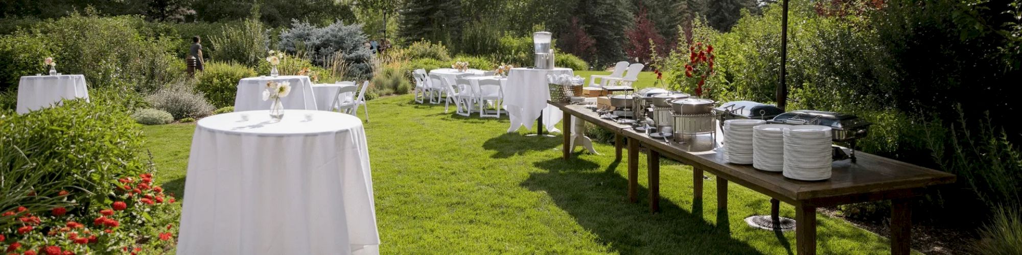 A garden setup with tables for dining, a buffet with a blue umbrella, surrounded by greenery and trees in the background. Perfect for an outdoor event.