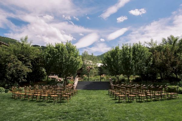 This image shows an outdoor setting with rows of chairs arranged for an event, possibly a wedding, surrounded by lush greenery under a blue sky.