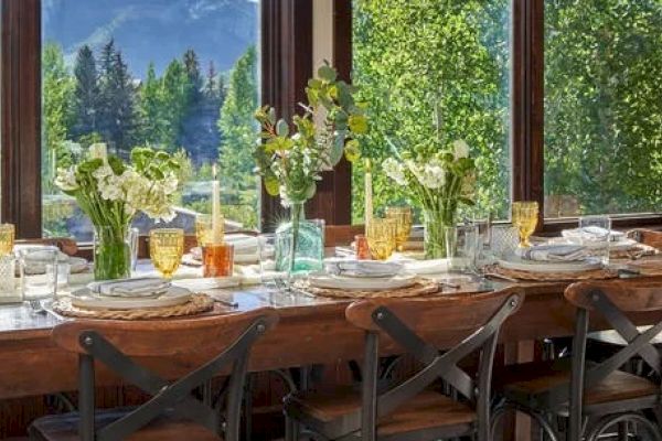 A dining table set for a meal, adorned with flowers and glassware, is situated in a room with large windows showing a scenic view of trees and mountains.