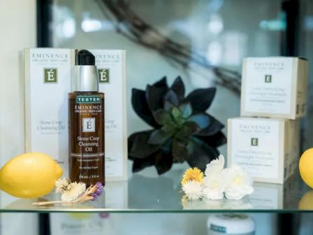 A display of Eminence Organic Skin Care products, including skincare boxes, a bottle of cleansing oil, lemons, flowers, and a succulent plant.