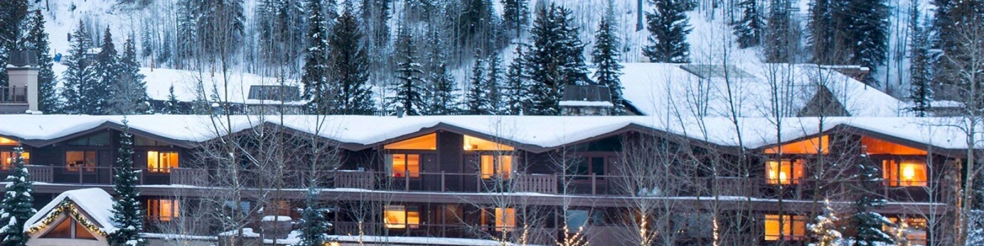 The image shows a cozy, illuminated mountain lodge nestled in a snowy landscape with pine trees and nearby ski slopes in the background.