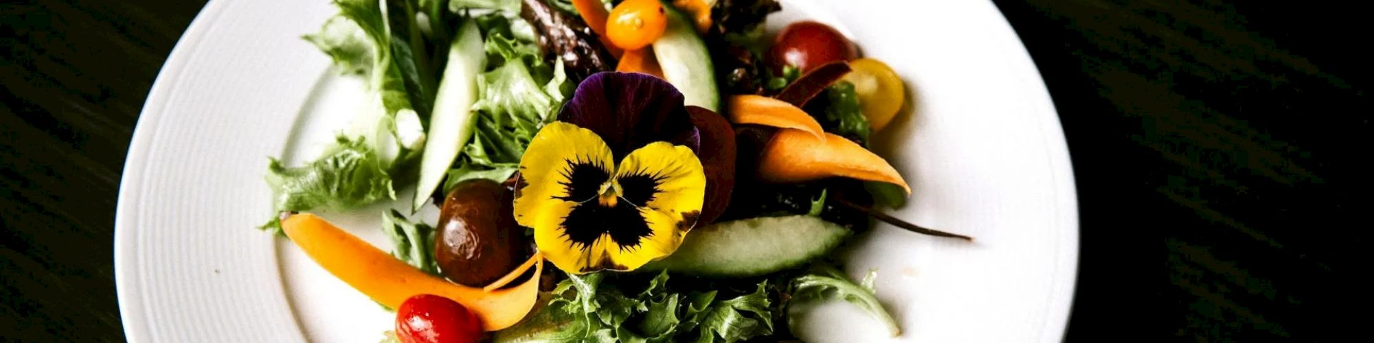 A fresh salad with mixed greens, cherry tomatoes, carrots, cucumber slices, and an edible flower on a white plate.