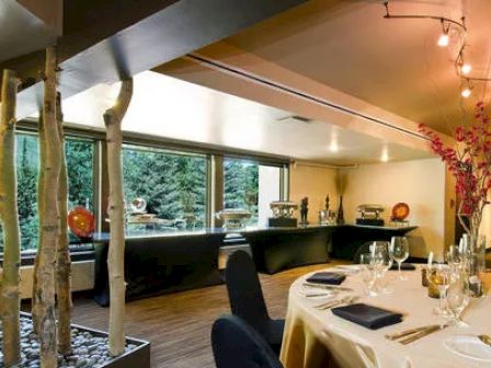 The image shows an elegant dining area with a set table, modern decor, unique lighting fixtures, and large windows providing a view of greenery outside.
