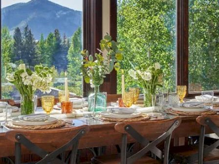 A dining table is set with plates, glasses, and flower arrangements, positioned by large windows offering a scenic view of trees and a distant mountain.