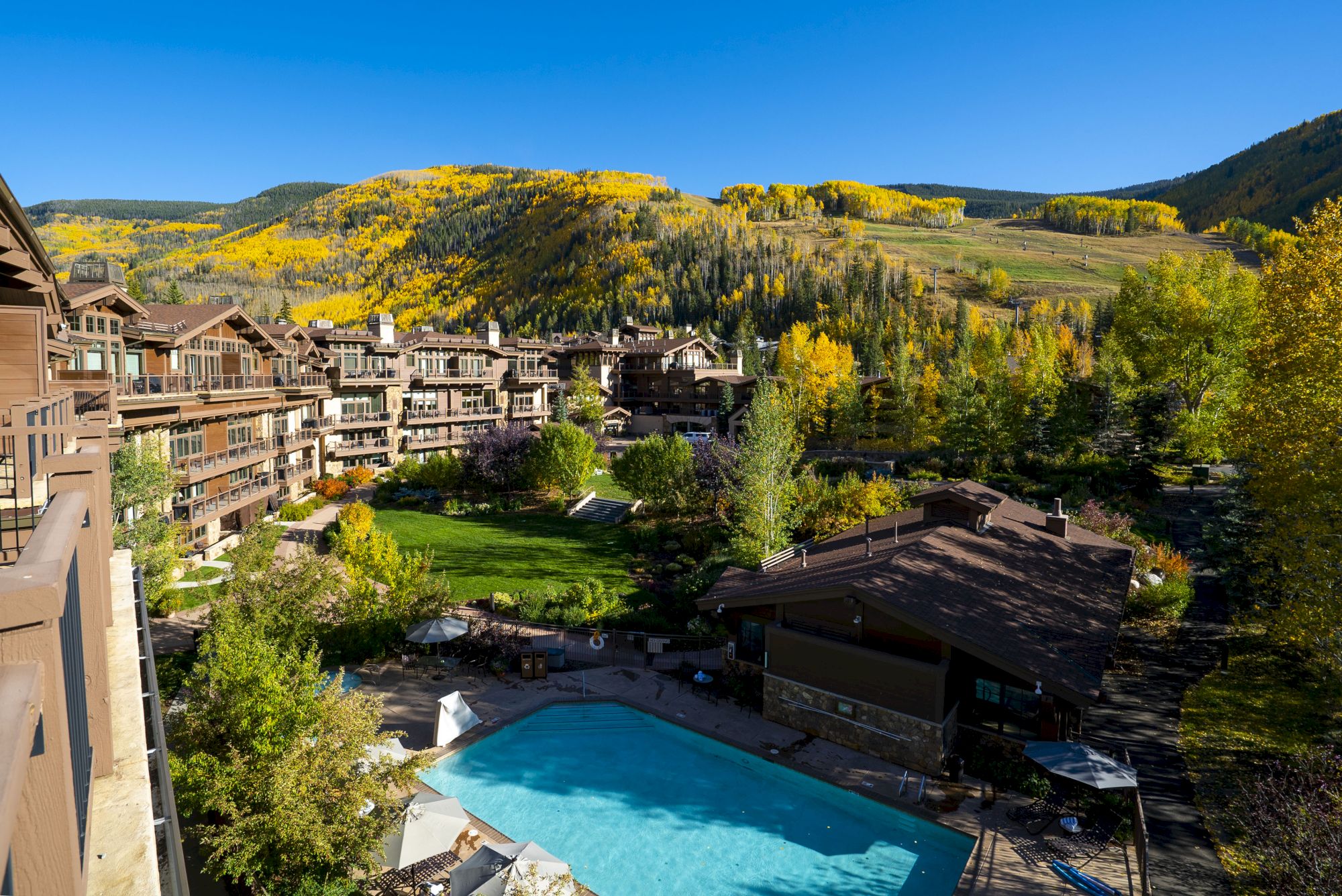 The image shows a resort with a pool, surrounded by buildings and lush hills with fall foliage under a clear blue sky.