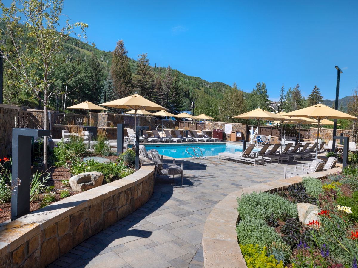 The image shows an outdoor pool area with lounge chairs, large umbrellas, and surrounding greenery, set against a backdrop of forested hills.