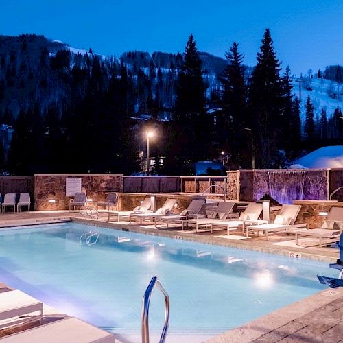 An outdoor pool area at dusk with lounge chairs, surrounded by a wooden fence and snow-covered mountains in the background.