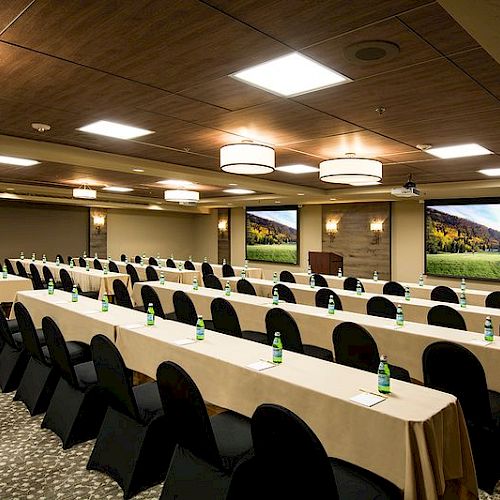 The image shows a modern conference room with rows of tables and chairs, a projector screen, and ambient lighting, ready for a meeting or seminar.