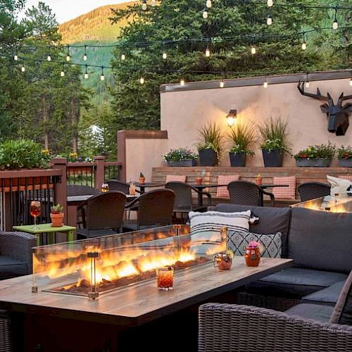 An outdoor seating area features a fire table, wicker furniture, string lights, and potted plants, creating a cozy atmosphere against a forest backdrop.