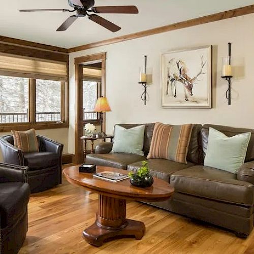 A cozy living room featuring a leather sofa, armchairs, wooden coffee table, deer artwork, and ceiling fan. Earthy tones add to the warm atmosphere.