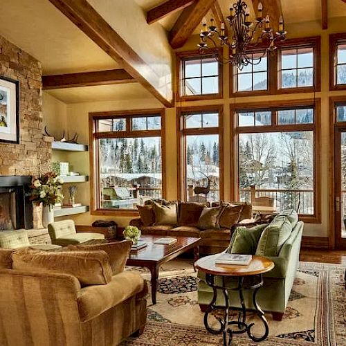 A cozy, rustic living room with large windows, a stone fireplace, plush sofas, a chandelier, and a scenic view of snowy mountains outside the windows.