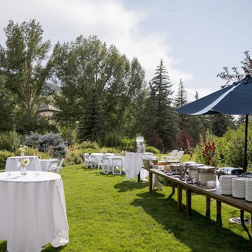 An outdoor event setup with round tables, white tablecloths, a buffet table with food covered under a blue umbrella, surrounded by greenery.