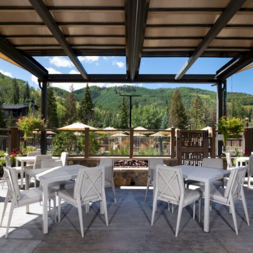 An outdoor patio with white tables and chairs under a pergola, overlooking a scenic view of trees and mountains with umbrellas in the background.