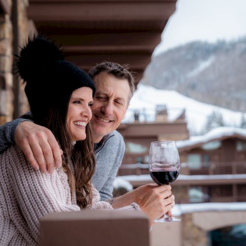 A couple dressed warmly, enjoying a drink on a balcony with a snowy mountain landscape in the background, smiling and looking content.