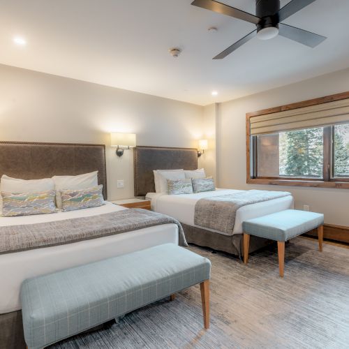 A modern hotel room features two queen beds with gray headboards, matching benches, wall sconces, a ceiling fan, and a window with a wooden frame.