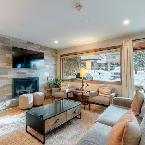 A cozy living room with a stone accent wall, fireplace, TV, and large windows overlooking a snowy exterior. Modern furniture completes the setup.