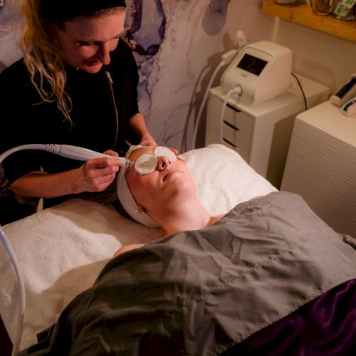 A person is receiving a facial treatment while lying on a bed, covered with a blanket, in a spa-like setting.