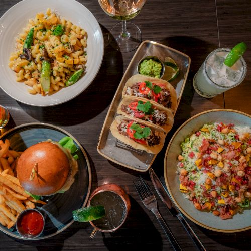 This image shows a spread of various dishes including pasta, tacos, a burger with fries, and a mixed salad, accompanied by drinks.