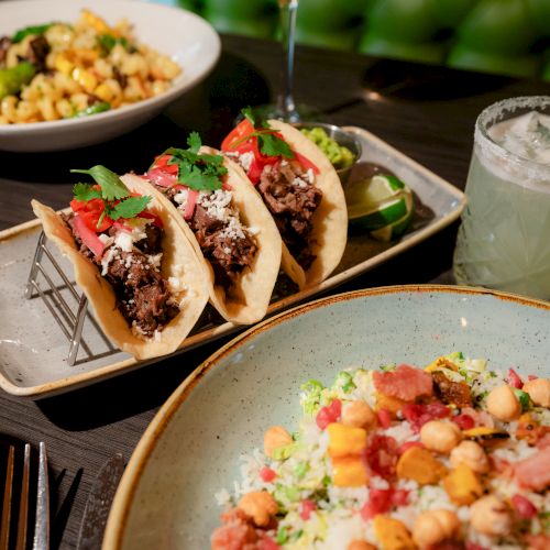 The image shows a plate of tacos, a dish of macaroni, a bowl of rice salad, and a cocktail with a lime wedge on top of it, all on a table.