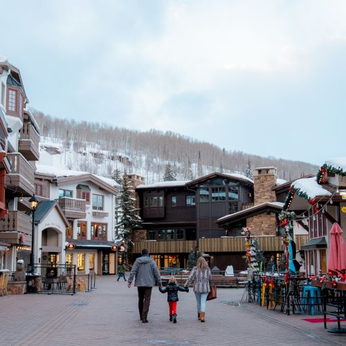 A family of three walks through a scenic snow-covered village, lined with charming buildings and decorated with festive holiday lights and garlands.