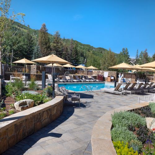 Outdoor pool area with lounge chairs, umbrellas, and trees in the background. Stone pathway and landscaped garden add to the scenic ambiance.