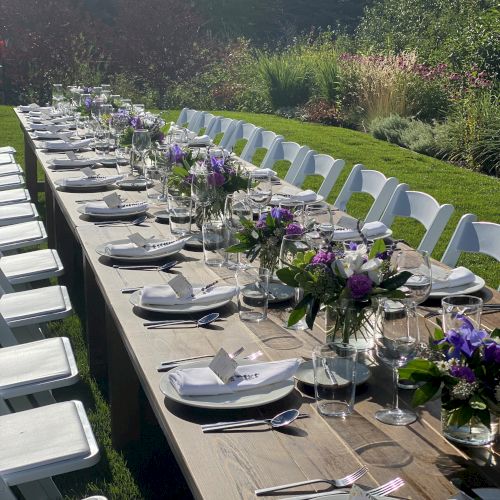 An outdoor event setup with a long table, white chairs, tableware, and floral decorations in a garden setting under sunlight, ready for guests.