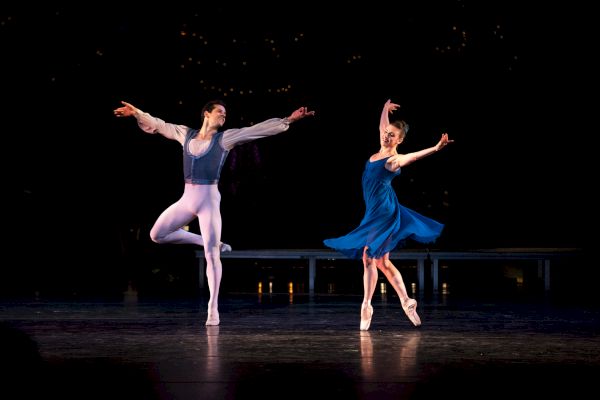 Two ballet dancers are performing on stage in elegant poses, with one in a blue dress and the other in white tights and a light vest.