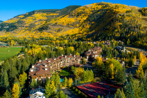 Aerial view of a mountain resort area with buildings surrounded by trees in autumn colors, set against a background of hills and blue sky.