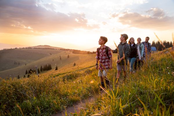 A group of people are hiking along a grassy, sunlit hillside with a scenic view and a colorful sunset in the background, walking along a path.