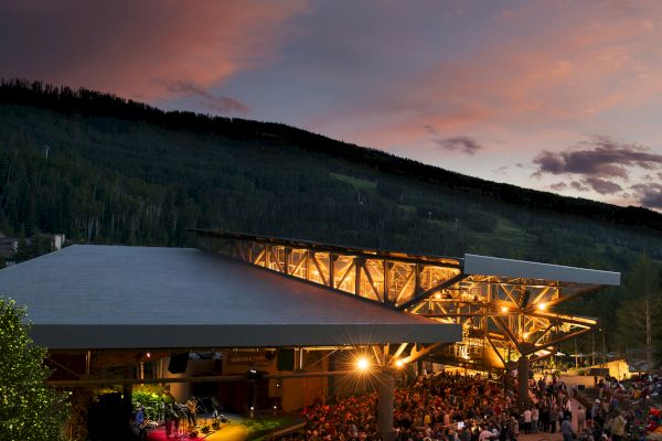 The image shows an outdoor concert venue at dusk, with a crowd gathered around the stage and a scenic mountainous backdrop.