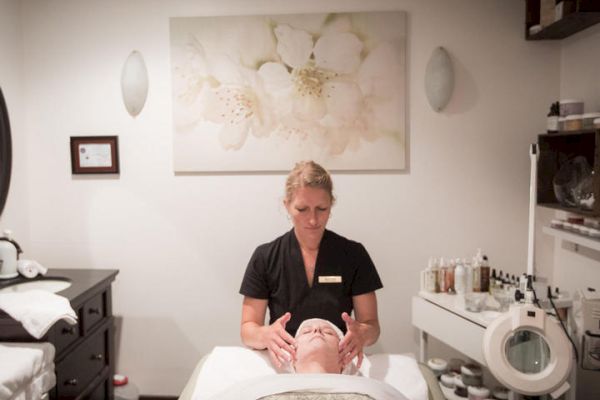 A woman receives a facial treatment from a therapist in a serene spa room decorated with floral art and skincare products on shelves.