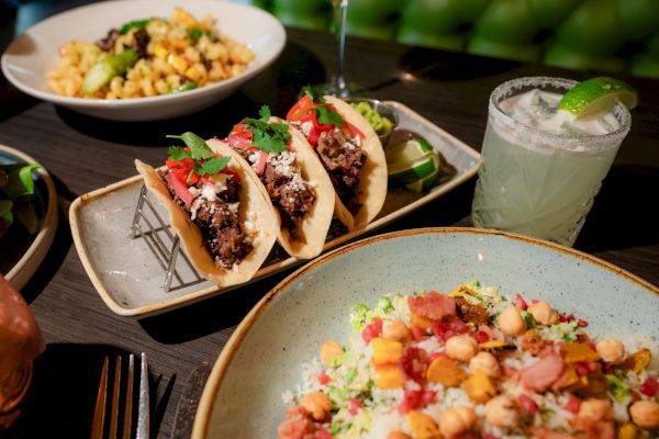 The image shows a table with beef tacos, a salad, a pasta dish, and a margarita garnished with a lime wedge, all on a dark surface.