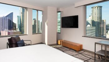 A modern bedroom with large windows offers a cityscape view, featuring a wall-mounted TV, chair, and a wooden bench.