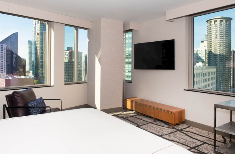 A modern bedroom with large windows offers a cityscape view, featuring a wall-mounted TV, chair, and a wooden bench.