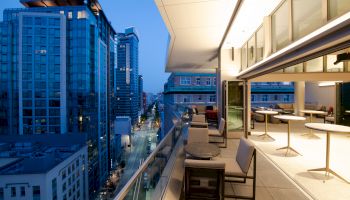 A modern rooftop balcony with seating overlooks a cityscape of tall buildings at dusk, creating an urban and sophisticated ambiance.