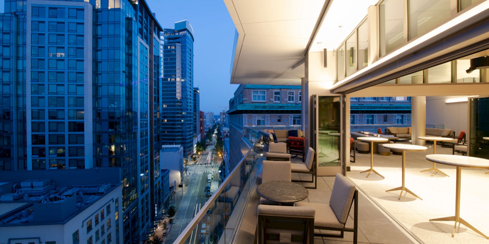 A modern cityscape viewed from a rooftop patio with tables and chairs, overlooking buildings and streets illuminated in the evening.