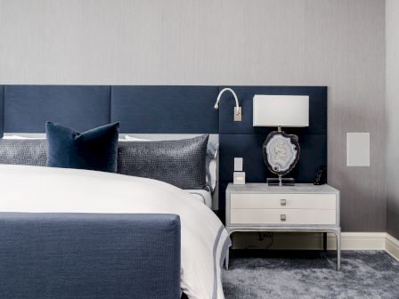 A modern bedroom features a blue upholstered bed, a white nightstand with a lamp and decorative piece, and gray carpet, creating a stylish ambiance.
