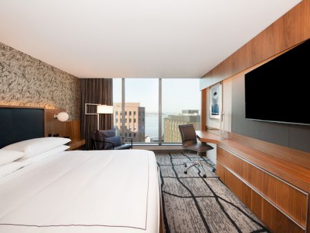 A modern hotel room with a large bed, wall-mounted TV, desk with chair, and a window offering a city skyline view.