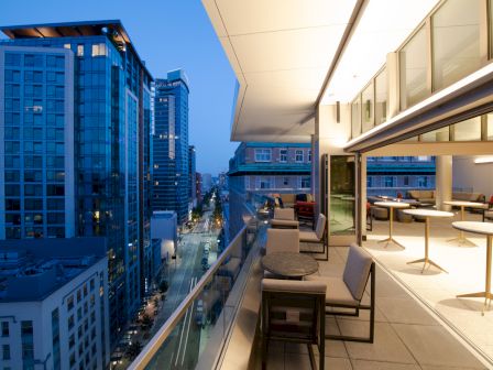 A modern cityscape at dusk, featuring tall buildings and a sleek apartment balcony with outdoor seating and tables, overlooking a bustling street.
