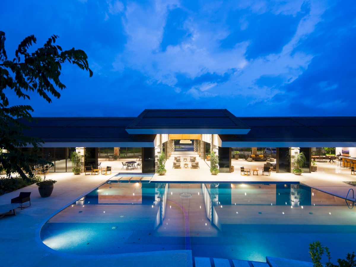 A modern building with an illuminated outdoor pool, surrounded by lounge chairs and greenery at dusk.
