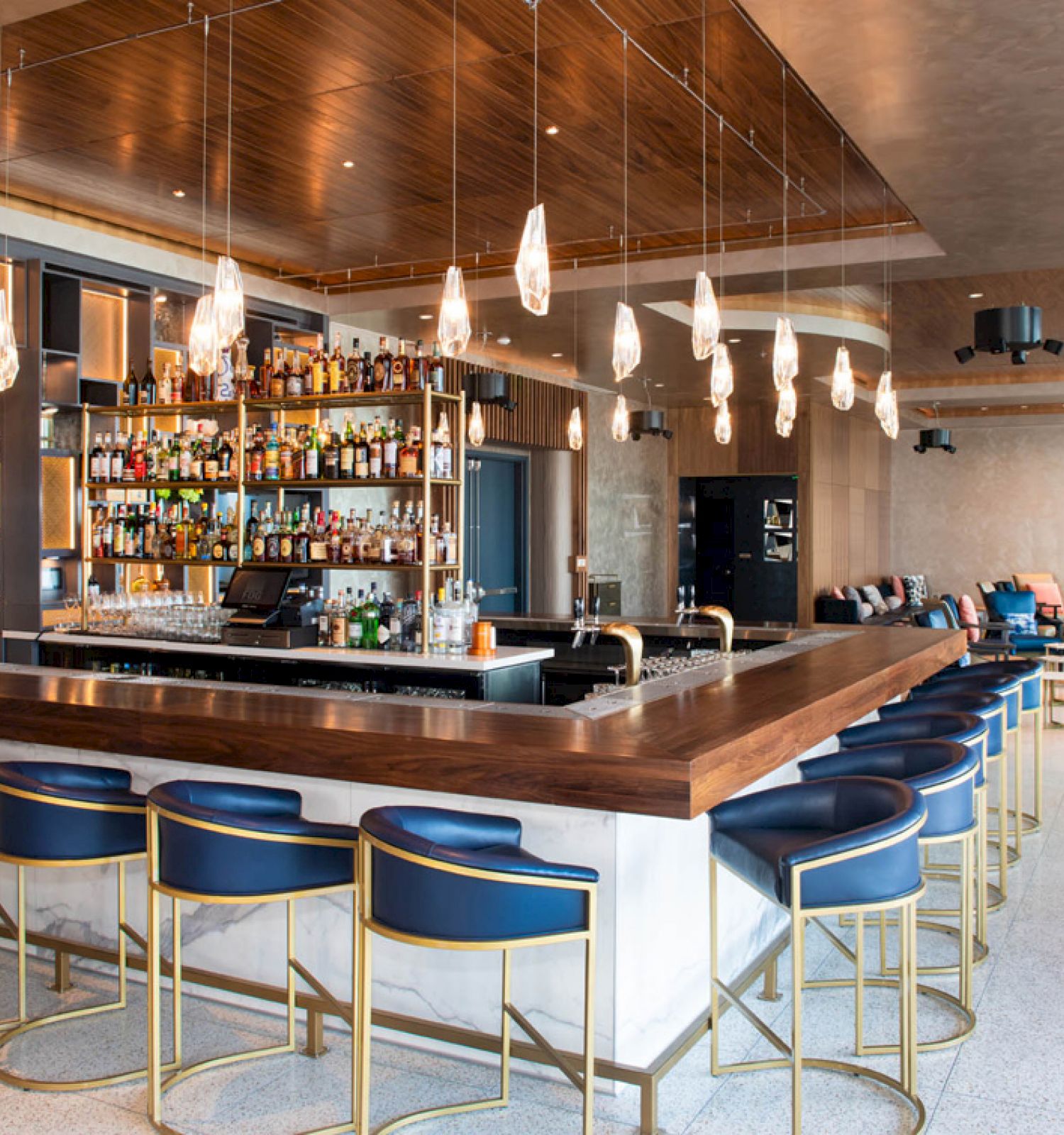 A modern bar with blue and gold bar stools, a stocked liquor shelf, hanging lights, and seating area in the background.